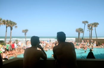 "Daytona" still with two men at the pool with the background of the beach and palm trees in front of them.