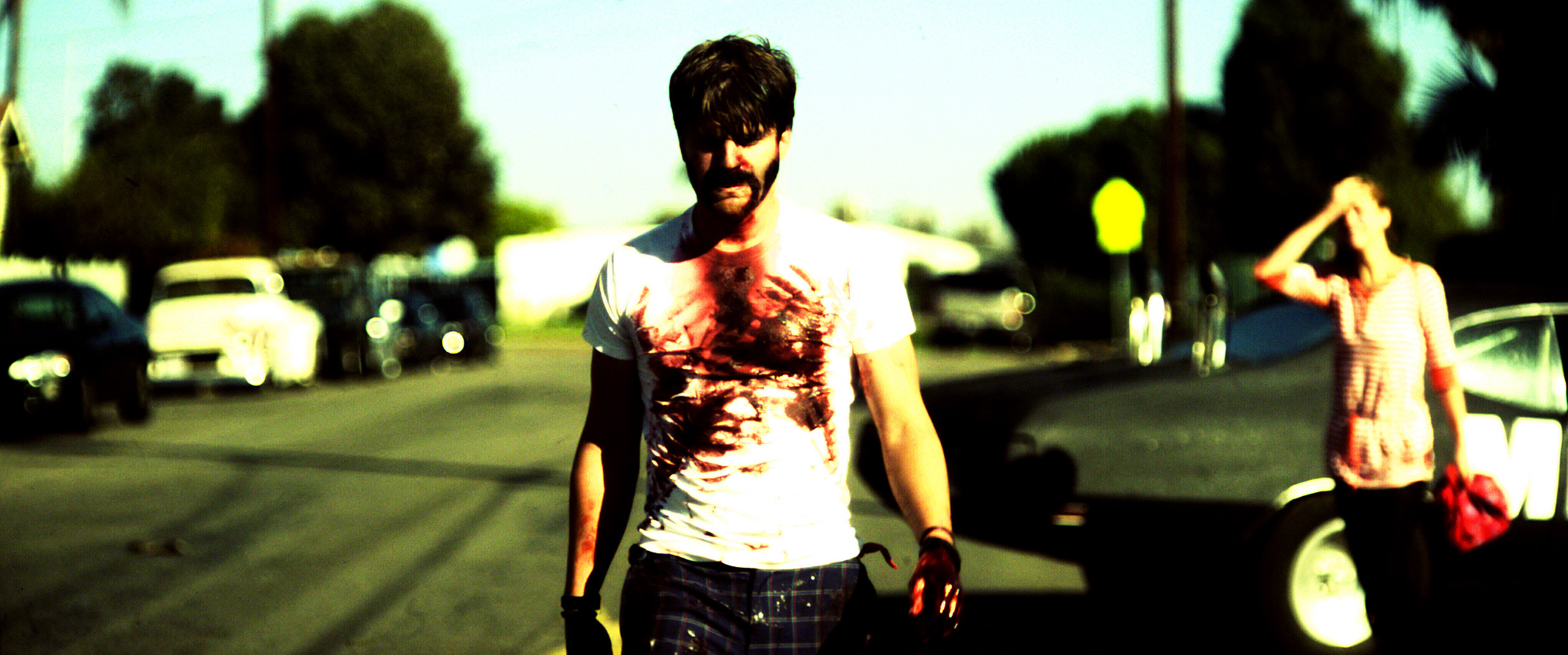 A still from Bellflower. A man with a bloodstained shirt walks down the street away from a woman looking on.