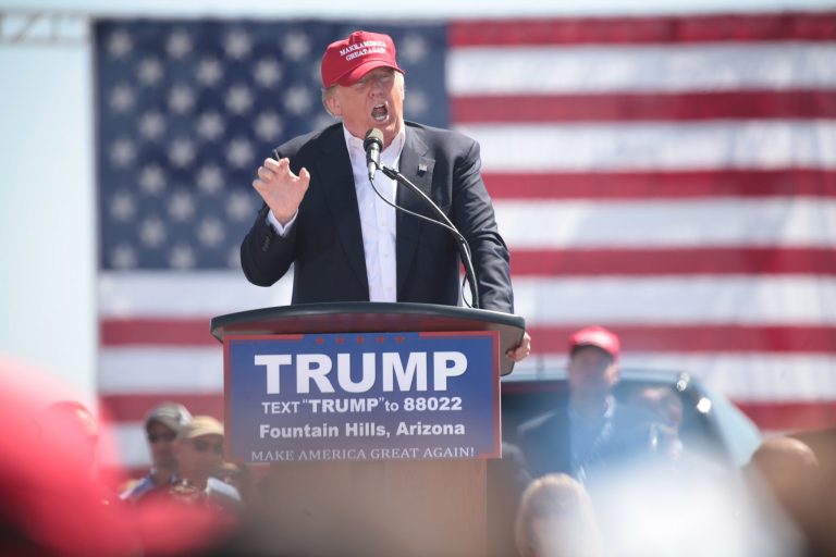 Donald Trump stands behind a podium, speaking in front of an American flag.