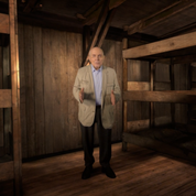 A bald man in black pants and a tan coat stands in a wooden barracks.