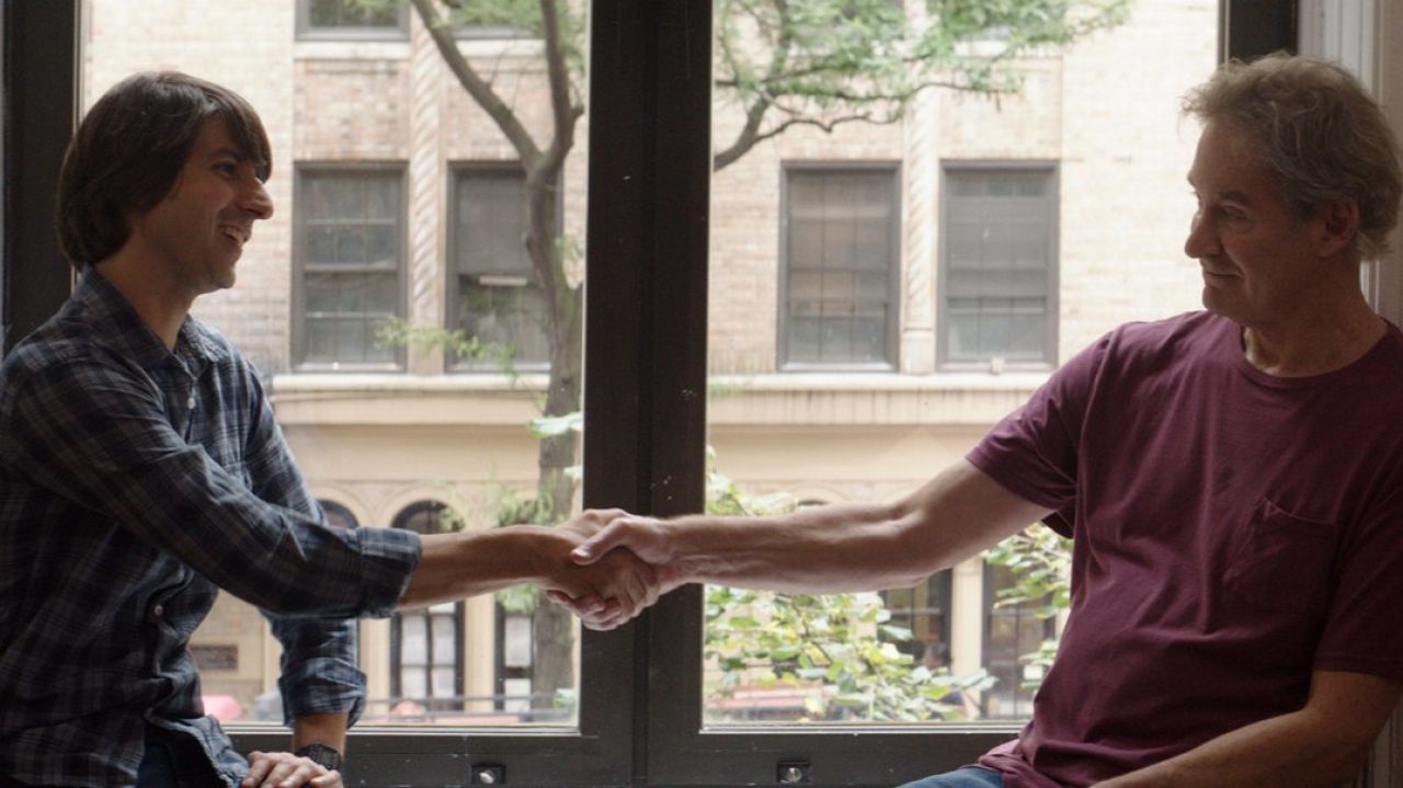 Demetri Martin's and Kevin Kline's shake hands in front of a large window in a still from the film.