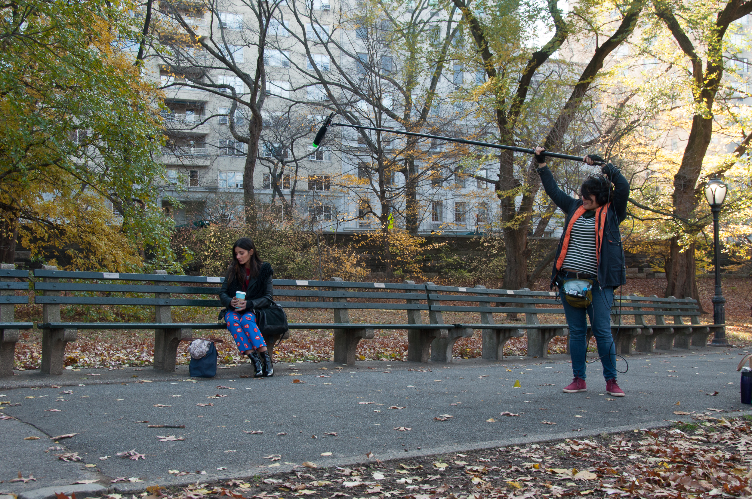 Sound designer holds boom microphone over actress who sits on a bench in a park