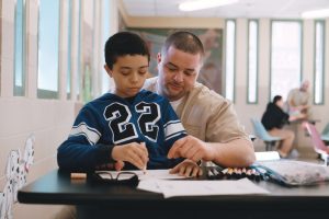 Maison sits on his father's lap coloring in the documentary Tre Maison Dasan.