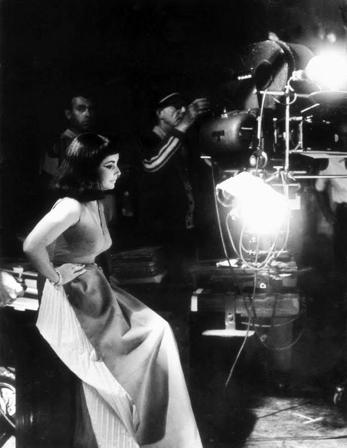 Elizabeth Taylor sits in a gown near a bright studio light.