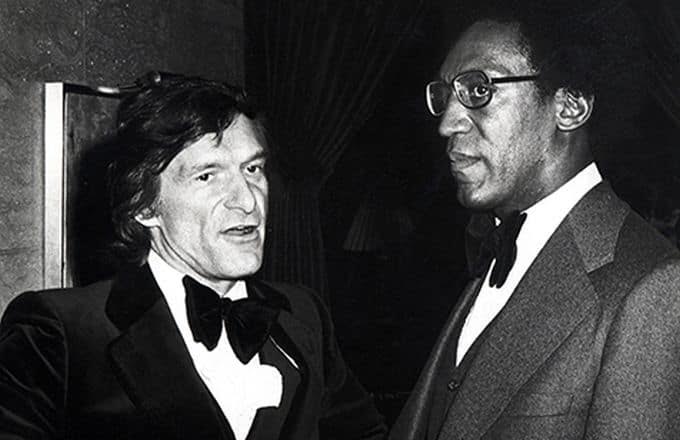 Hugh Hefner and Bill Cosby are wearing suits and speaking to one another.
