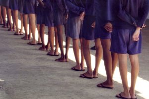 A still from the Maria Ramos documentary Behave that shows minors standing in a line from behind at a juvenile detention facility where you can just see their torsos, legs, and feet in matching blue shorts and sandals.