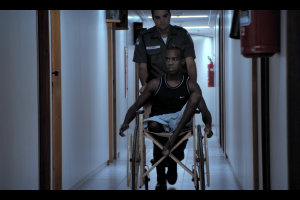 A police officer wheels a disabled man in a wheelchair to his court date in the Maria Ramos documentary Justice.