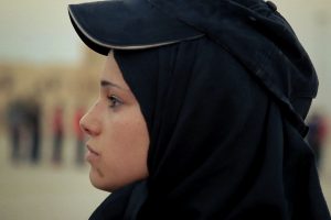 A still from the documentary What Walaa Wants, which is a close up of her face of her wearing a hijab and a hat looking toward the left paying attention to someone offscreen.