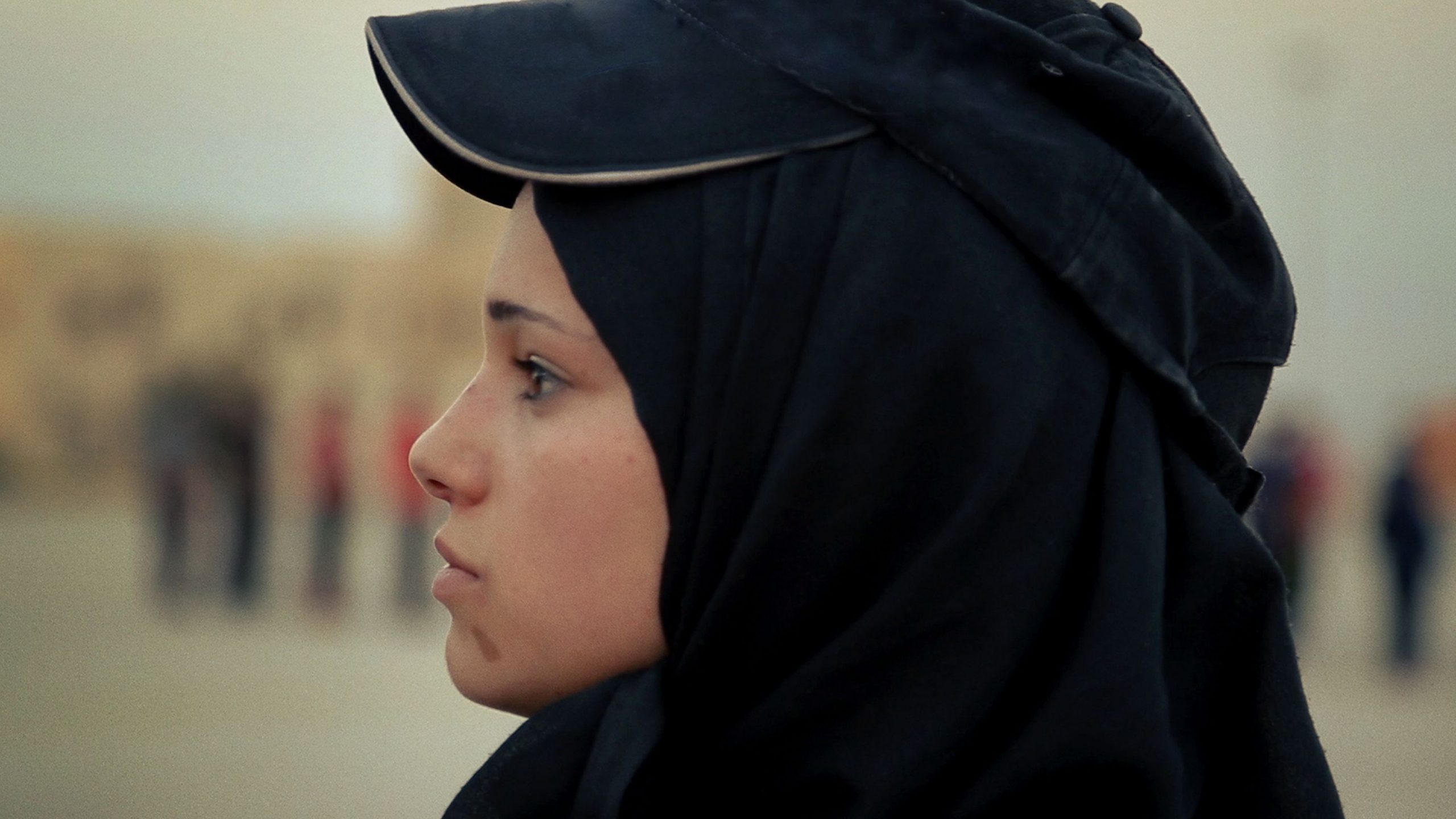 A still from the documentary What Walaa Wants, which is a close up of her face of her wearing a hijab and a hat looking toward the left paying attention to someone offscreen.