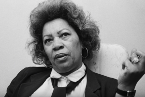 A black and white photo of Toni Morrison from when she was editor at Random House looking sideways at the camera and holding up her clenched fist 