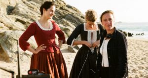 Two actresses in perdio dress stand on the beach as the director pauses the scene. 