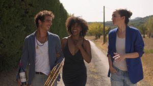 A man and woman, linking arms, walk down a dirt path with a second woman. All three are smiling. 