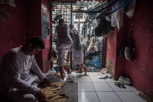 Men lean on the walls and bars of a small prison cell. 