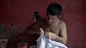 A shirtless man with tattoos sews. 