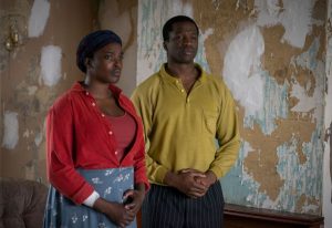 A man and woman stand inside a room with wall paper peeling. 