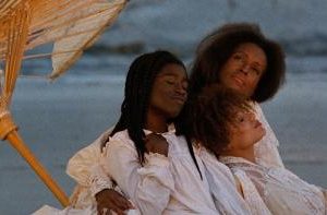 Three women in white dresses wrapped closely to each other under an umbrella on the beach
