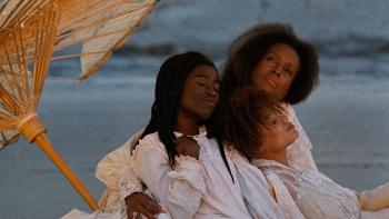 Three women in white dresses wrapped closely to each other under an umbrella on the beach